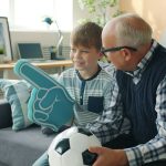 Joyful family grandfather and grandson are watching football on TV cheering hugging celebrating victory. Sports fans, modern lifestyle and leisure concept.